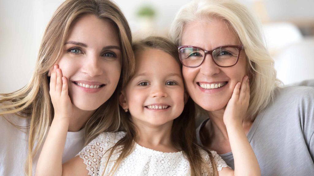 A young girl, with a bright smile, surrounded by her mother and grandmother, all sharing a big smile